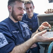 uniformed brown-haired and bearded engineering technician weapons engineering working with 3D Printed part smiling auburn haired colleague in background