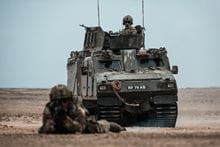 A Royal Marine in the desert in foreground lying on the floor with a gun pointing at the camera and a vehicle in the background.
