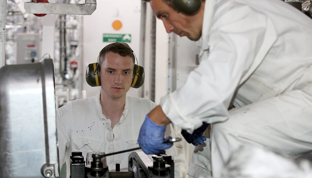 royal-navy-careers-RFA-cadetships-08 Two navy personnel work whilst wearing protective clothing and ear defenders.