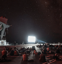 Service people on a flight deck on a ship watching a big screen