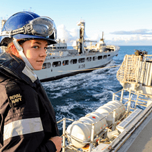 A person in a blue helmet stands on a ship at sea and looks back slightly towards the camera. Another ship is in the background.