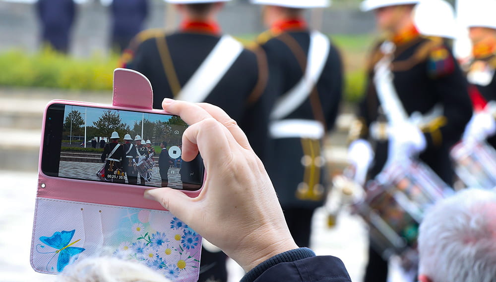 Royal-Navy-Families-08-Social-Media Grey-haired woman holding up a digital camera with marching band displayed the screen