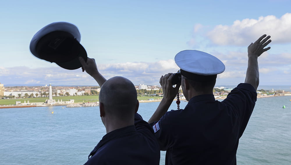 Royal-Navy-Families-10-Keeping-in-Touch Two sailors on boat looking to shore with back to camera, one waving hat
