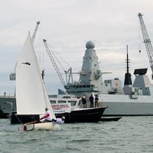 A small white dingy and harbour patrol boat sail in front of a destroyer