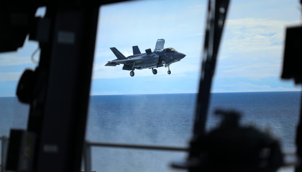 An image of an aircraft flying with a view from inside the bridge of a ship