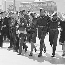 A black-and-white photo shows British and US Service personnel celebrating with Londoners in Piccadilly Circus.