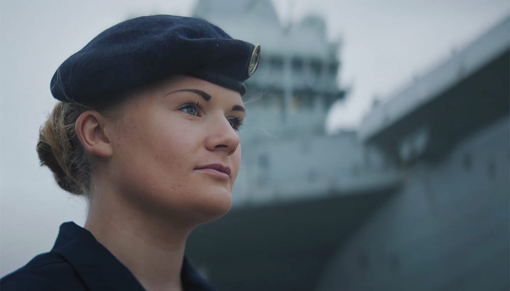 uniformed royal navy sailor with aircraft carrier in background looking into distance