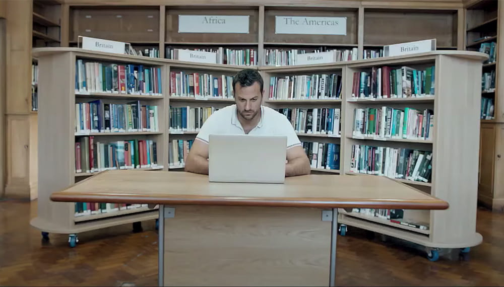 brown haired white t-shirted person sits behind laptop with bookshelves behind
