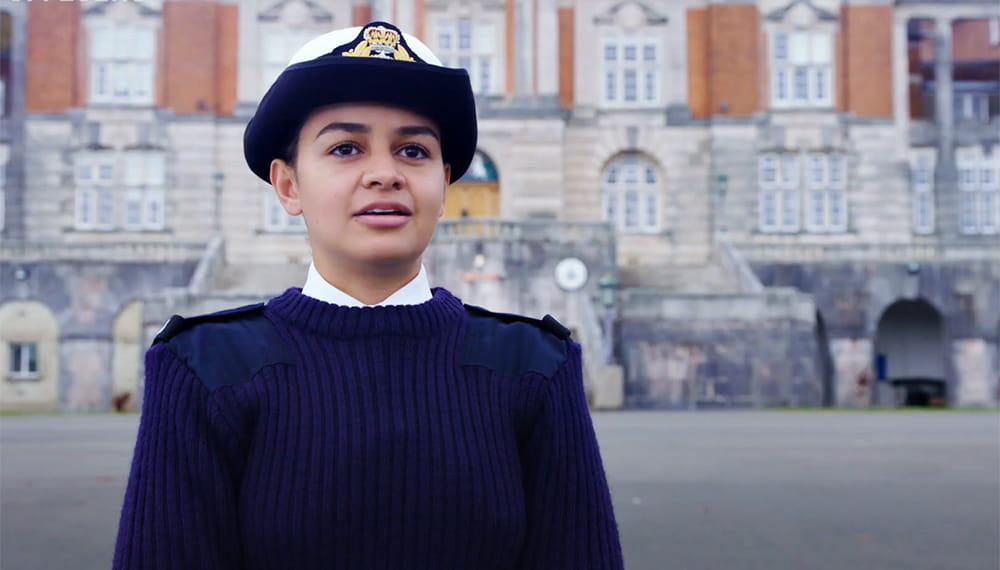 officer cadet wearing blue jumper and officers white hat looks into distance with brnc, britannia royal naval college, in background