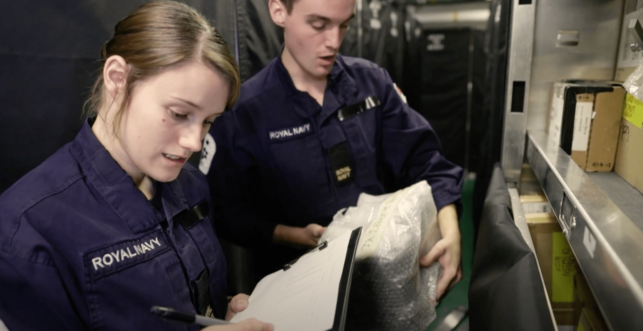Two supply chain logisticians onboard a ship working together to sort stock, one is writing on a clipbaord and the other is reading out a label from an item of stock