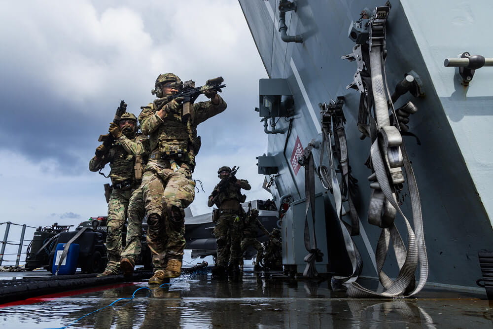 A colour photo shows several Royal Marines on the deck of a ship. The ground is noticeably wet and skies are grey. The Commandos wear full fatigues and hold automatic weapons in hand. They stealthily approach a corner with guns aimed. In the background, 