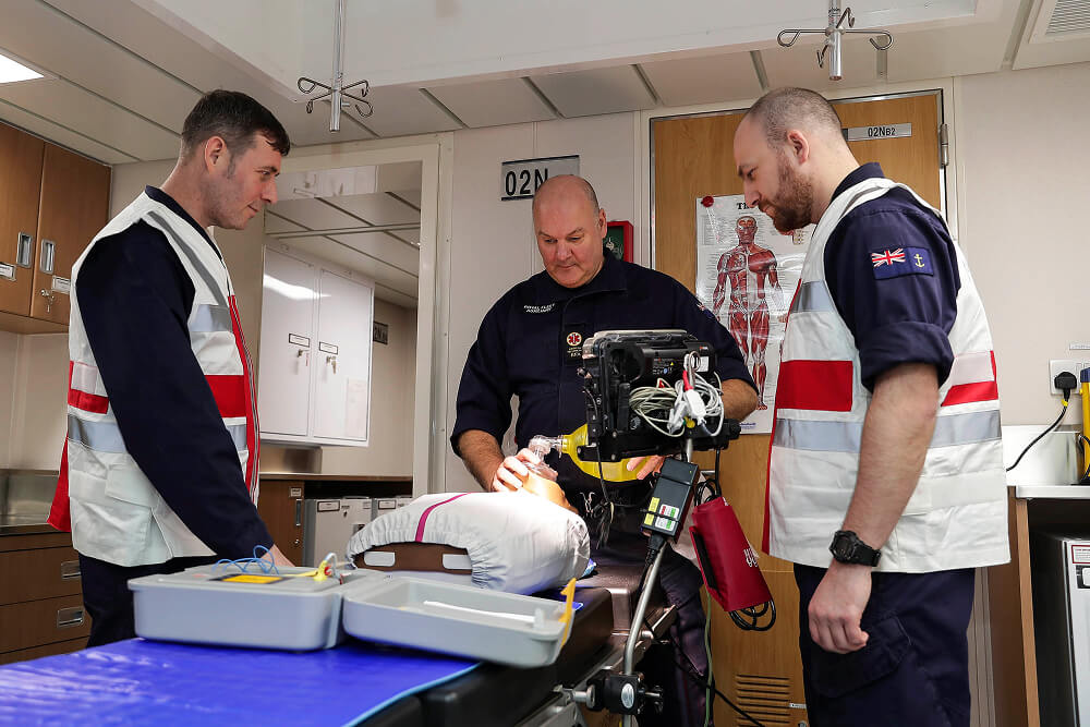 A colour photo shows an RFA medical technician demonstrating the use of specialist equipment for two trainees. In the centre is a table with a dummy and an open kit. A small monitor stands just to the side. The technician holds a breathing apparatus in his hands and wears a blue Royal Navy work uniform. The two trainees look on intently and wear blue Royal Navy work uniforms and protective medical vests. 