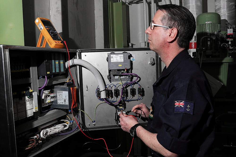 A colour photo shows an RFA engineering technician working on an electrical panel. There are several components and wires running across the panel. He observes readings on an external monitoring device that is hooked up to the panel. He wears a blue Royal Navy work uniform.