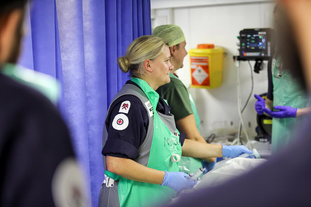 A colour image shows an over-the-shoulder perspective of a Royal Navy medical assistant near a patient's bedside. The bed and patient are just out of frame. The medical assistant is wearing a blue Royal Navy work uniform, sterile gloves, and a sterile protective vest. In her hand is a syringe. In the background are two more medical personnel in conversation.