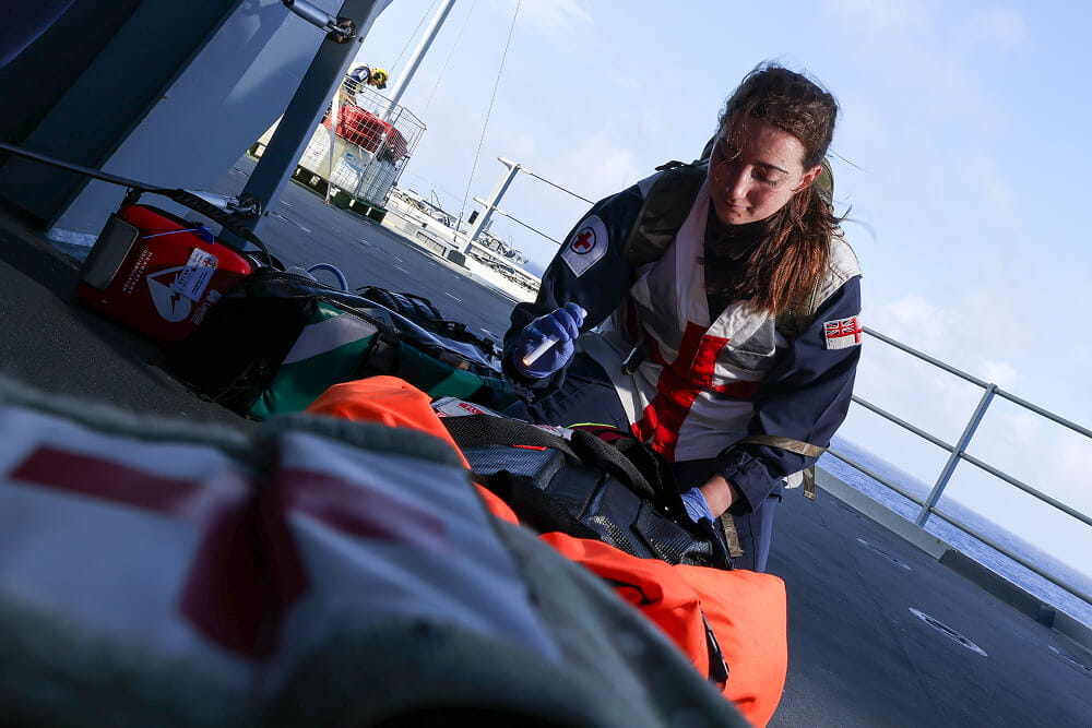 A colour image shows a Royal Navy medical officer kneeling down on a ship's deck. She is wearing a blue Royal Navy work uniform, a protective vest, and a utility backpack. She examines several medical supply bags with a flashlight in hand.