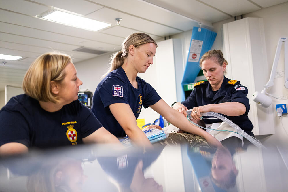 A colour image shows two Royal Navy medical students and a medical officer attending to a patient. The patient and bed are just out of frame. The medical officer places a medical tube onto breathing apparatus, while the students hold the patient and observe. The students wear blue Royal Navy work shirts with medical insignias, while the officer wears a blue Royal Navy work uniform. 