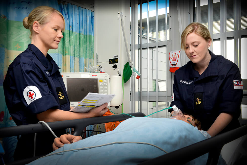 A colour image shows two Royal Navy medical servicemembers attending to a patient in a bed. They both wear blue Royal Navy work uniform. The patient lies flat on their back with a breathing appartus attached to their face. In the background are medical devices,  monitors, and a privacy curtain.