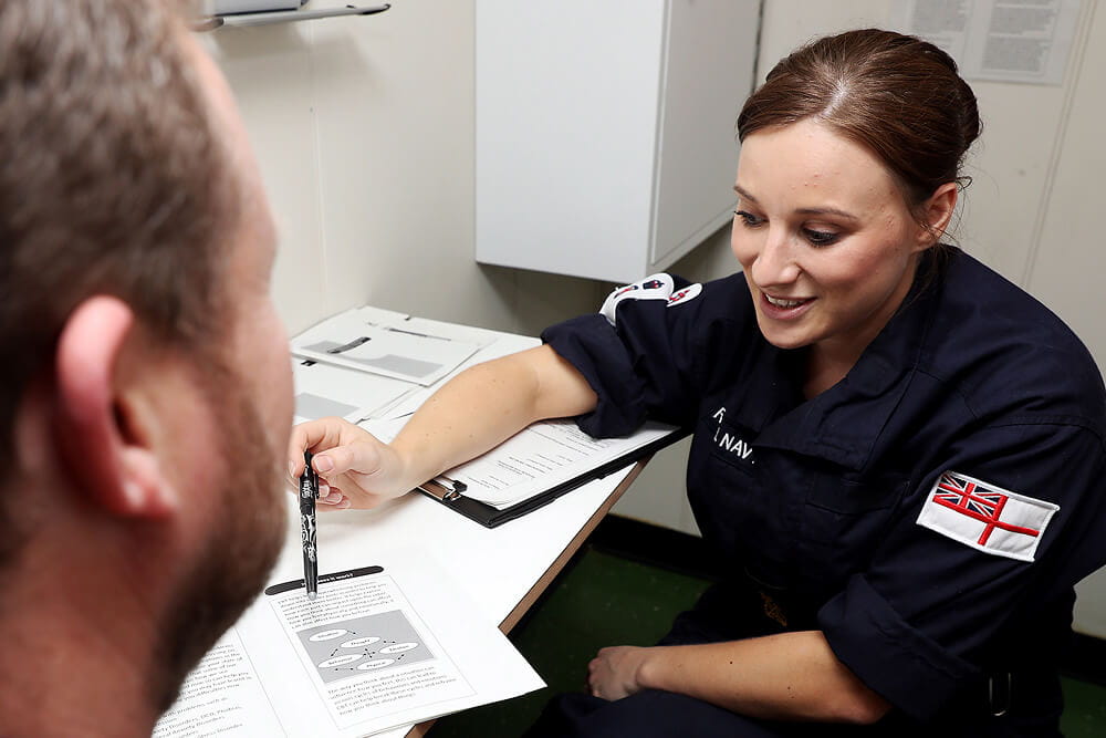 A colour image shows a Royal Navy nurse sitting at a desk with a clipboard and several printed sheets. She wears a blue Royal Navy work uniform. She is chatting with a patient and drawing attention to an informational diagram in one of her print-outs. The image is cropped and the patient's side profile is visible.