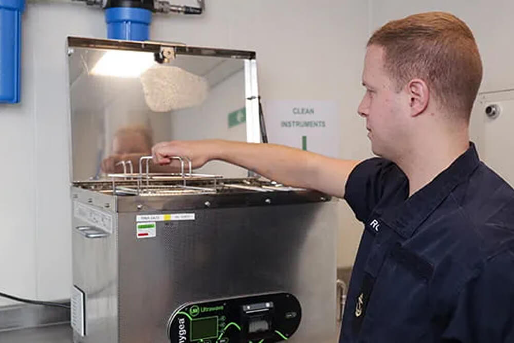 A colour images shows a Royal Navy operating department practicioner operating a large machine for disinfecting medical instruments. He wears a blue Royal Navy work uniform.