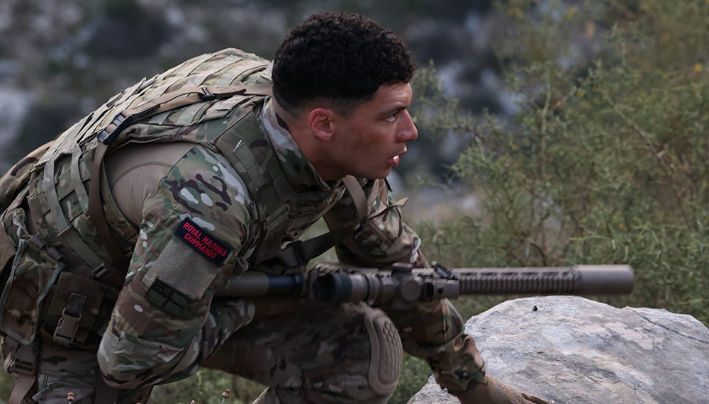 Member of Royal Marines wearing full equipment and holding a weapon looking forward. Camouflaged amongst plants and rocks. 