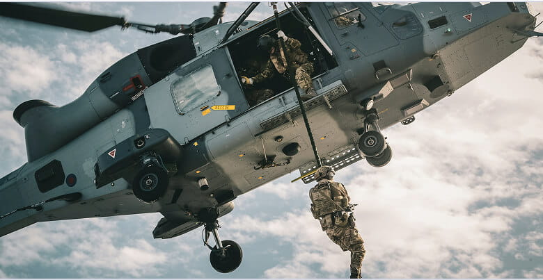 A Royal Marine is dangling from a helicopter on a cord with another Royal Marine assisting them.