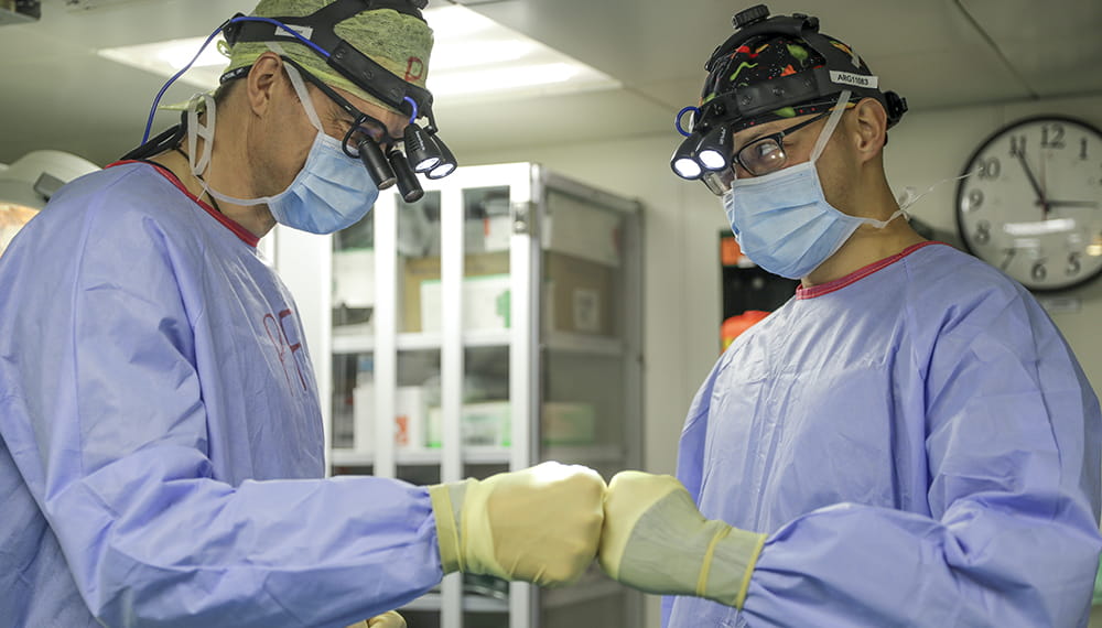 medics in light blue theatre robes with ocular headsets and yellow protective gloves fist bumping in clinical setting