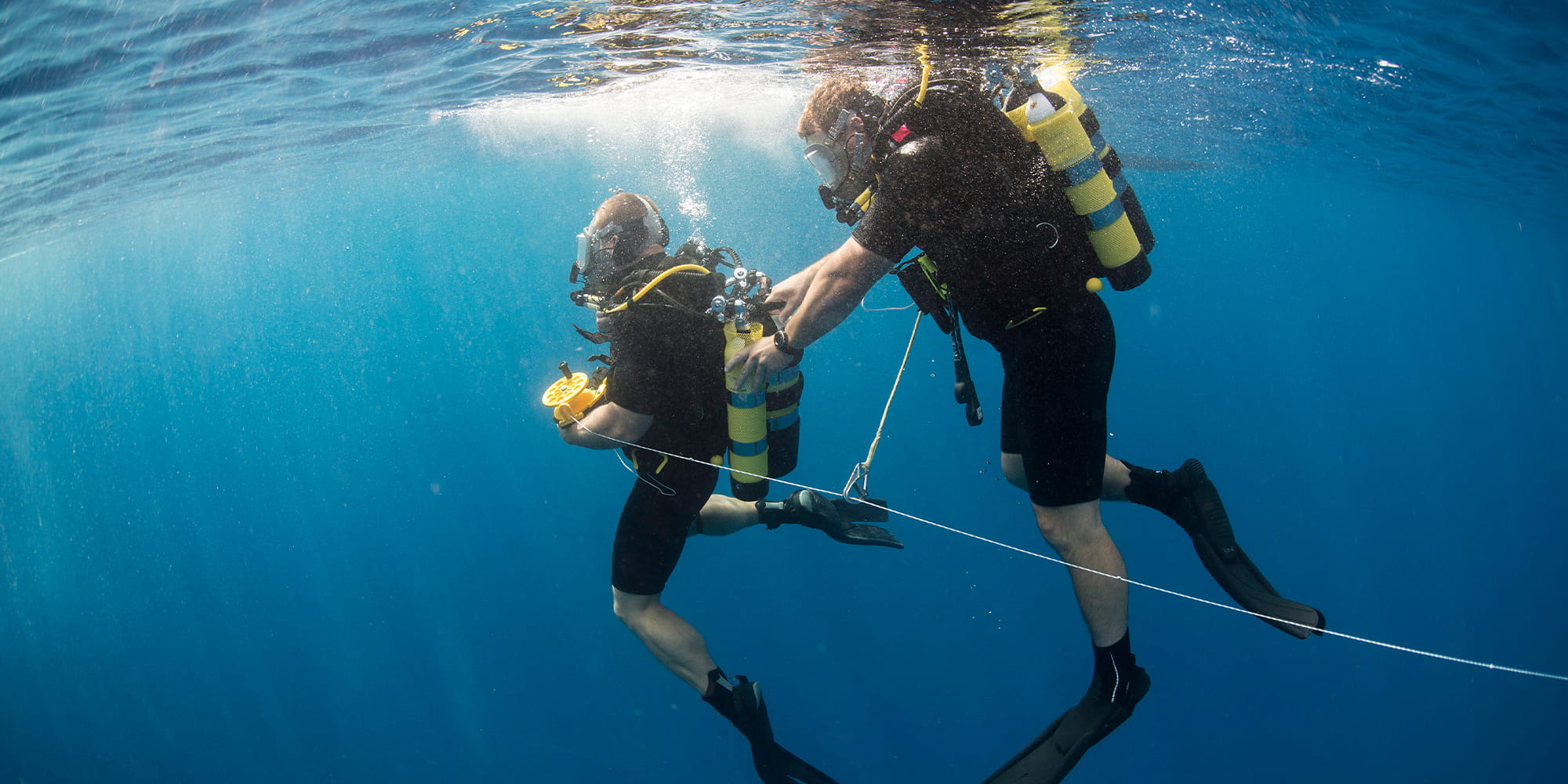 2 divers under the surface of the sea, one adjusts the others oxygen tank